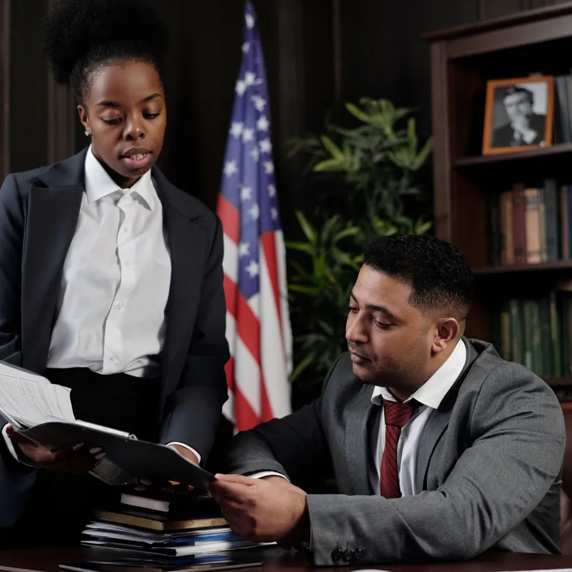 Lawyer at a desk reviewing a file handed over by a colleague in an office with a US flag and bookshelves