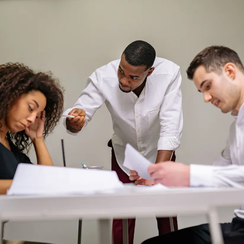 Frustrated team at a meeting table, one colleague leaning in and pointing a pen while others read papers