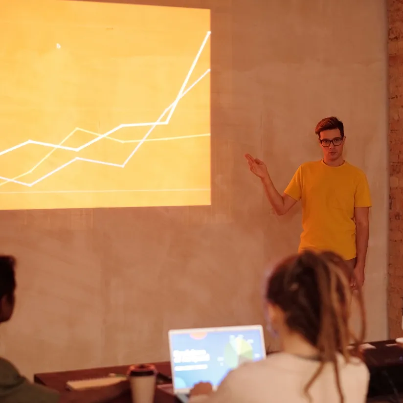Presenter in a yellow T-shirt pointing to a projected rising line chart as colleagues watch with laptops