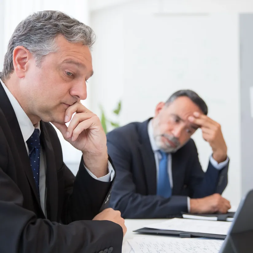 Two concerned executives in suits at a meeting table, one with his hand on his chin, one rubbing his forehead