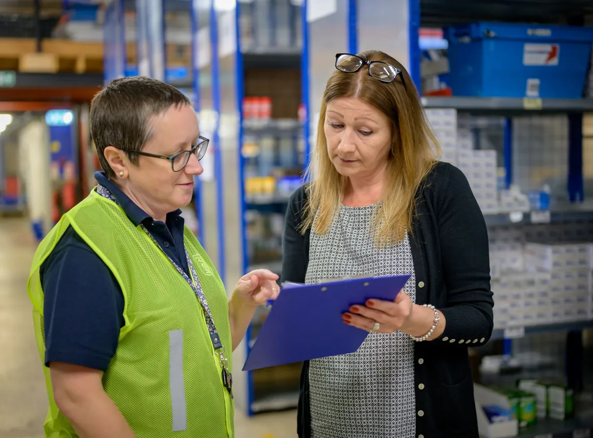 Warehouse supervisor in a hi-vis vest reviewing a clipboard with a colleague between stocked shelves