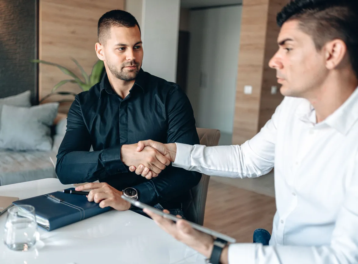 Two businessmen shaking hands at a white meeting table, one holding a tablet beside a notebook