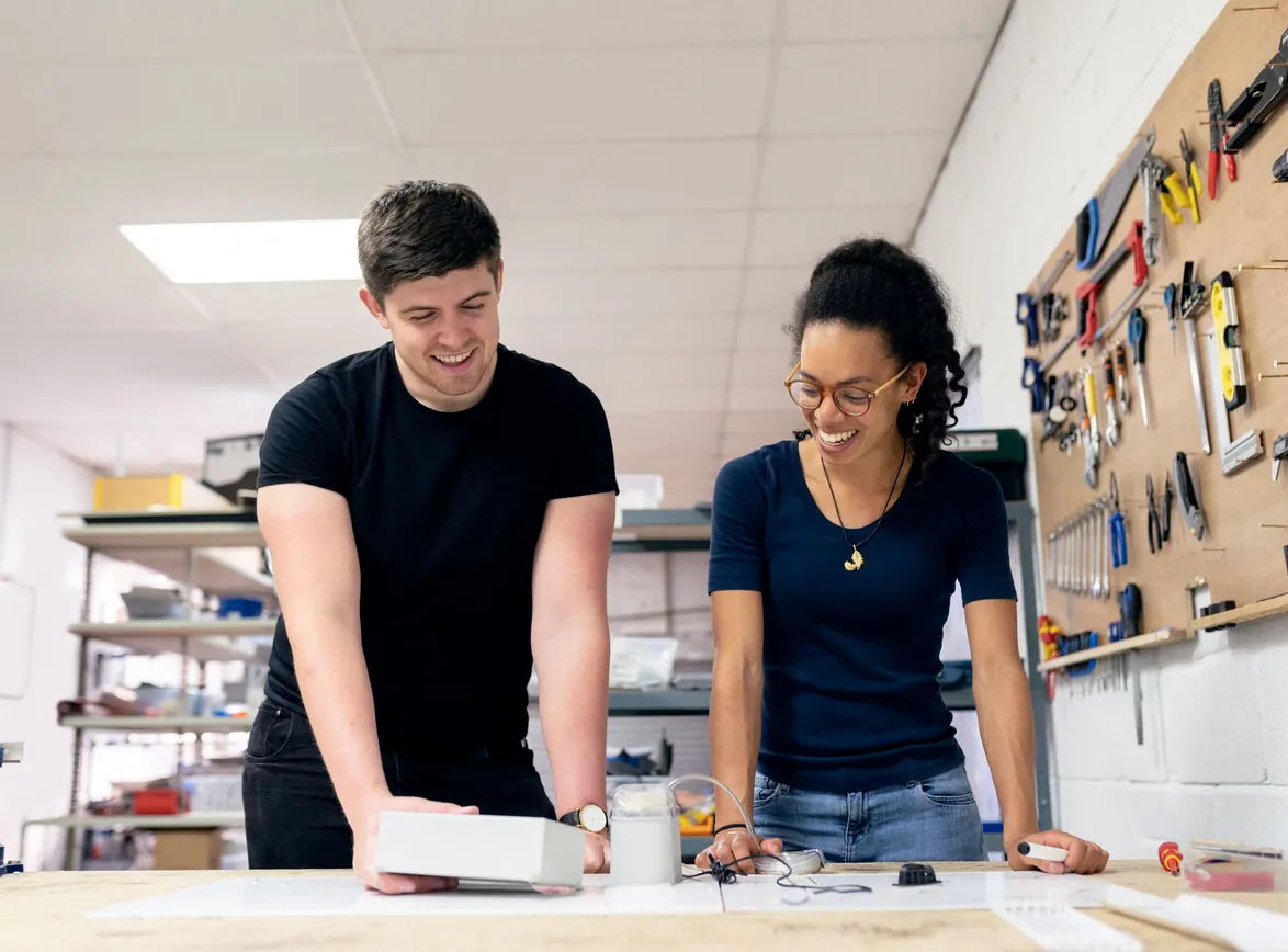 Two colleagues smiling while working on a small device at a workbench in a workshop with tools on a pegboard