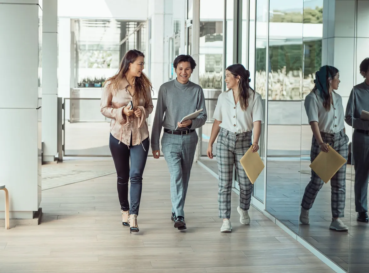 Three colleagues walking and talking together along a bright glass-walled office corridor