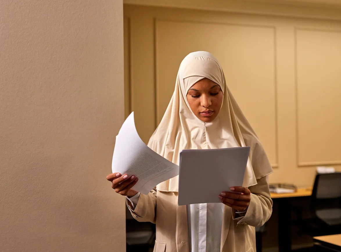 Woman in a hijab and beige suit reading through printed documents in an office