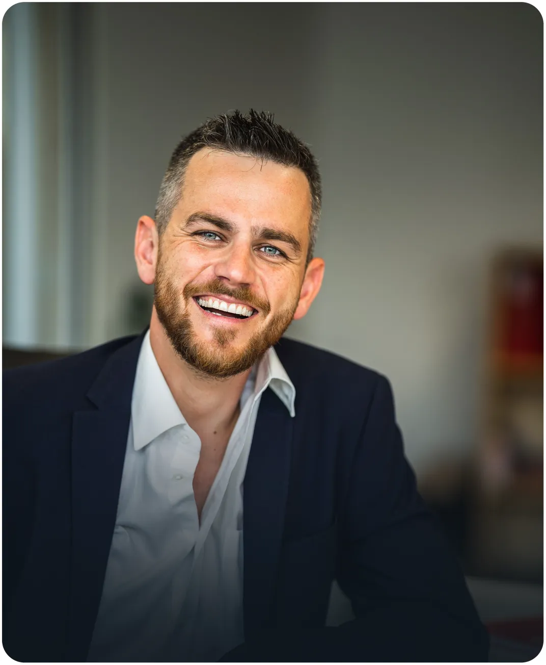 Smiling bearded businessman in a navy blazer and open-collar white shirt seated in an office