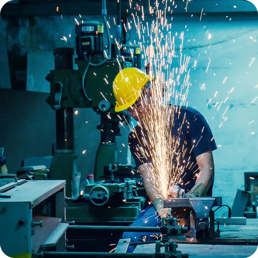 Factory worker in a yellow hard hat cutting metal with an angle grinder as sparks fly in a machine shop