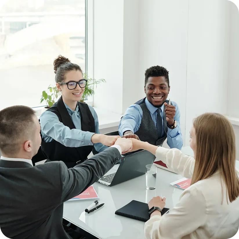 Four professionals bumping fists over a conference table with a laptop, notebooks and printed brochures