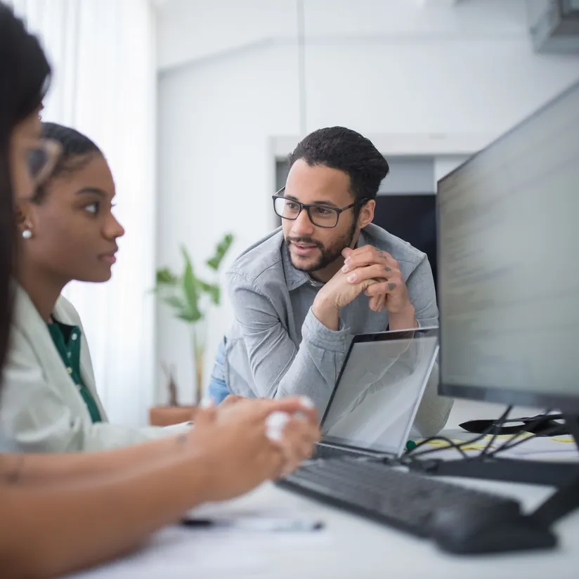 Software developers talk at a shared workstation with a monitor, keyboard and laptop in a bright office