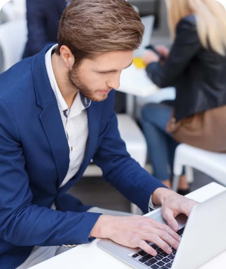 Professional in a navy blazer typing on a laptop at a white table, with a colleague on her phone behind him