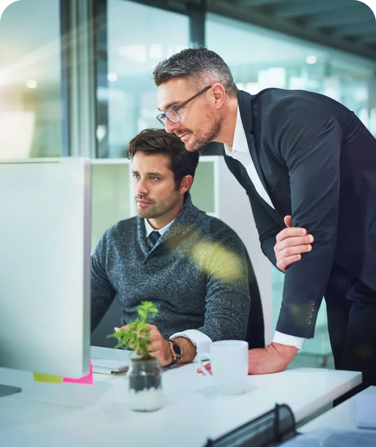 Manager in glasses leaning over a seated colleague's shoulder as they both look at a desktop monitor