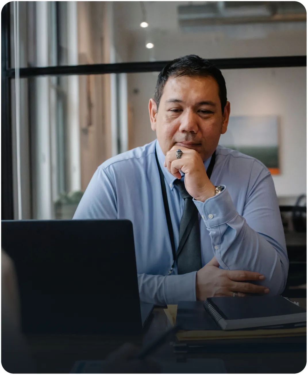 Executive in a blue shirt and tie, chin on hand, reviewing a laptop beside a stack of notebooks