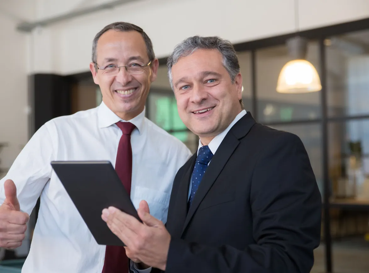 Two smiling businessmen in corporate attire discussing work on a tablet in a modern office