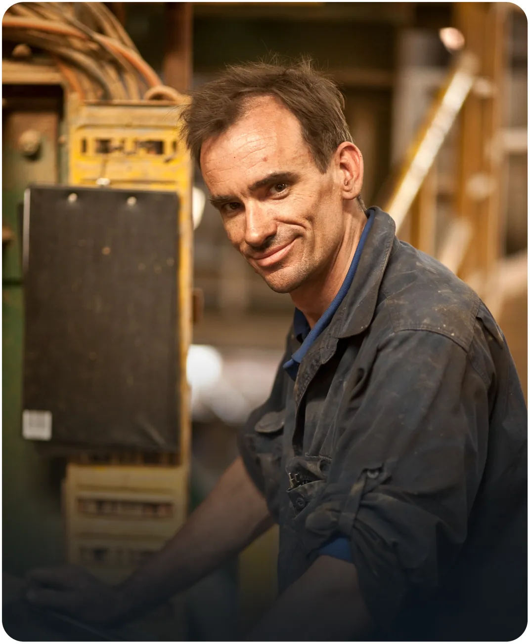 Factory technician in a worn work shirt smiling beside an industrial machine control panel