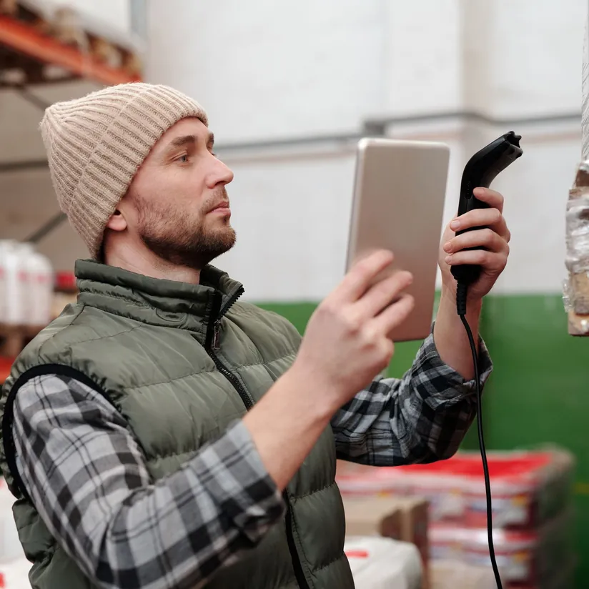 Warehouse worker holding a tablet and handheld barcode scanner while checking wrapped pallets on storage shelves