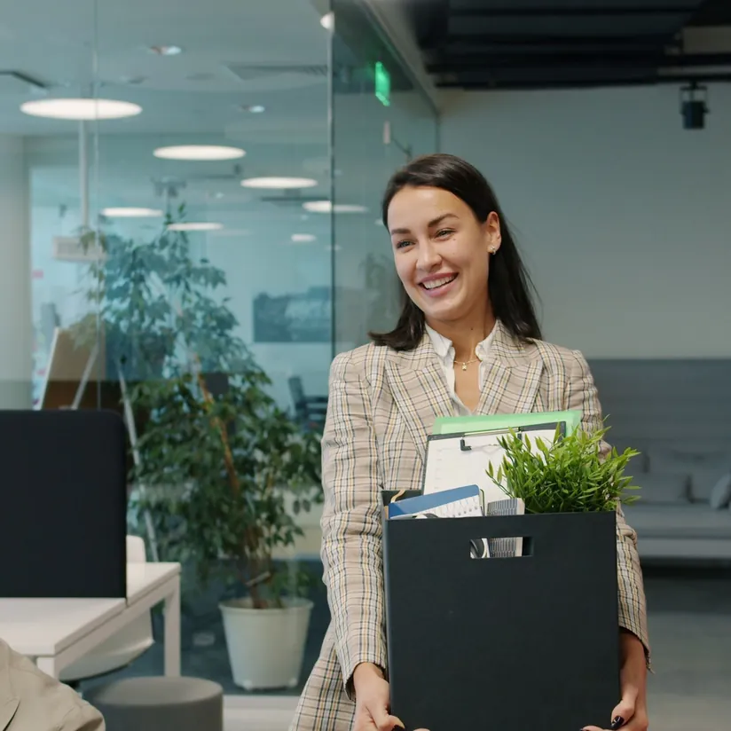 Smiling employee carrying a box of folders, a clipboard and a potted plant through a modern office