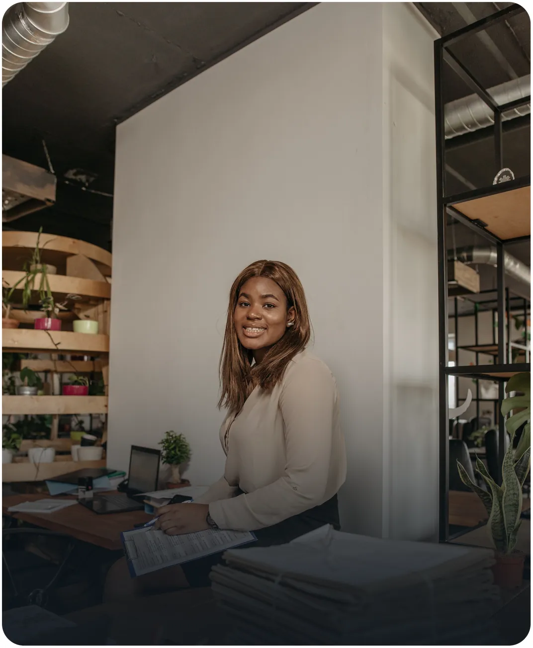 Smiling office worker holding a clipboard with a form beside stacks of paperwork in a loft-style office