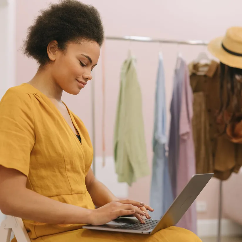 Woman in a mustard dress typing on a laptop in front of a clothing rack of hanging garments