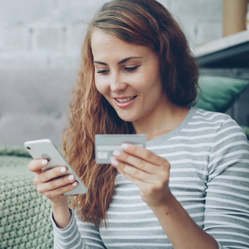 Smiling woman holding a smartphone and a payment card while shopping online at home