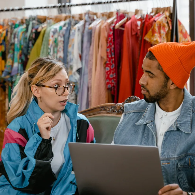 Two colleagues talking over a laptop in front of racks of colorful vintage clothing