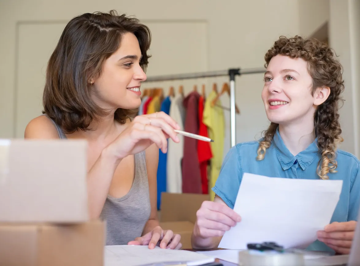 Two businesswomen discussing ideas in an office setting with clothing in the background