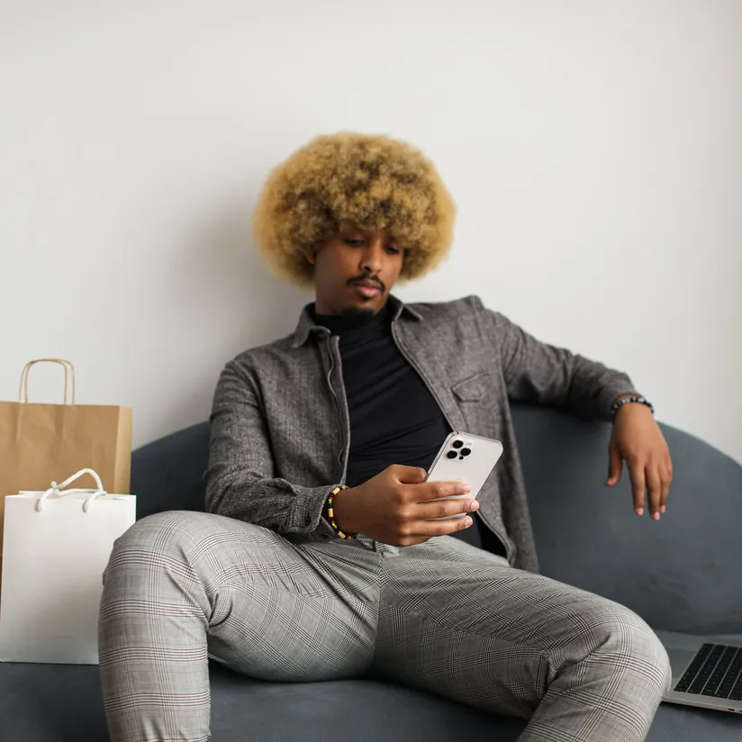 Man on a sofa browsing on his smartphone beside paper shopping bags, with a laptop next to him