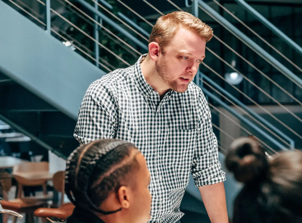 Advisor in a checked shirt leans in to talk with seated team members in a modern office
