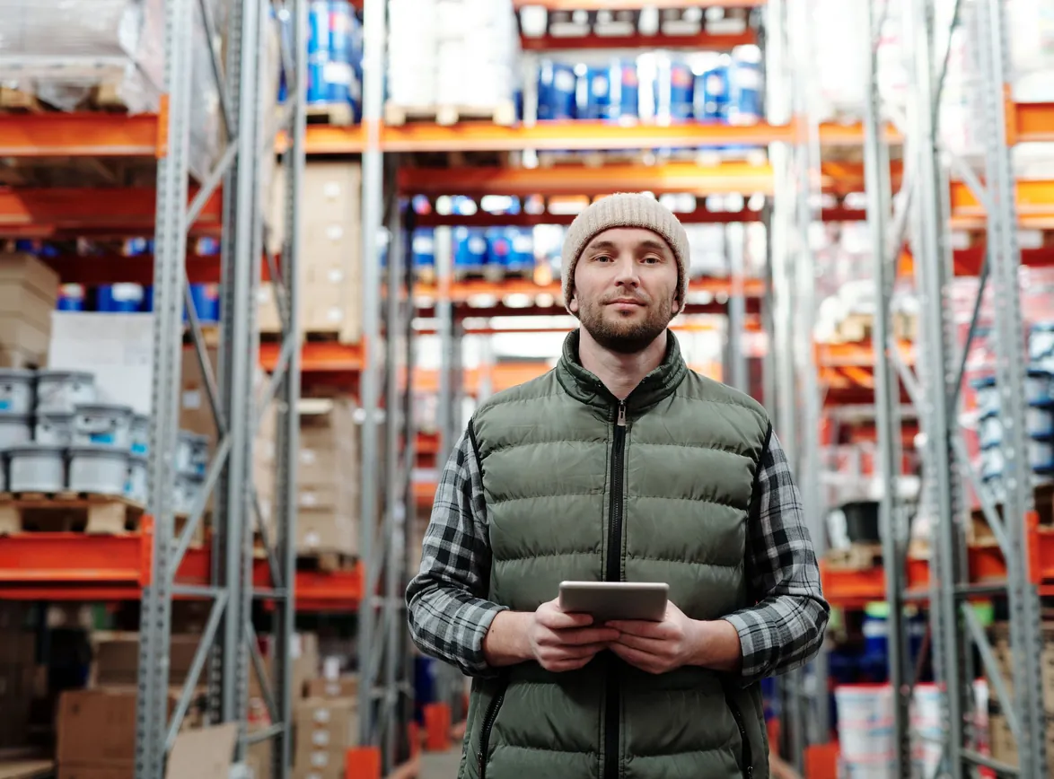 Warehouse manager in a beanie and padded vest holds a tablet in an aisle of stocked pallet racks