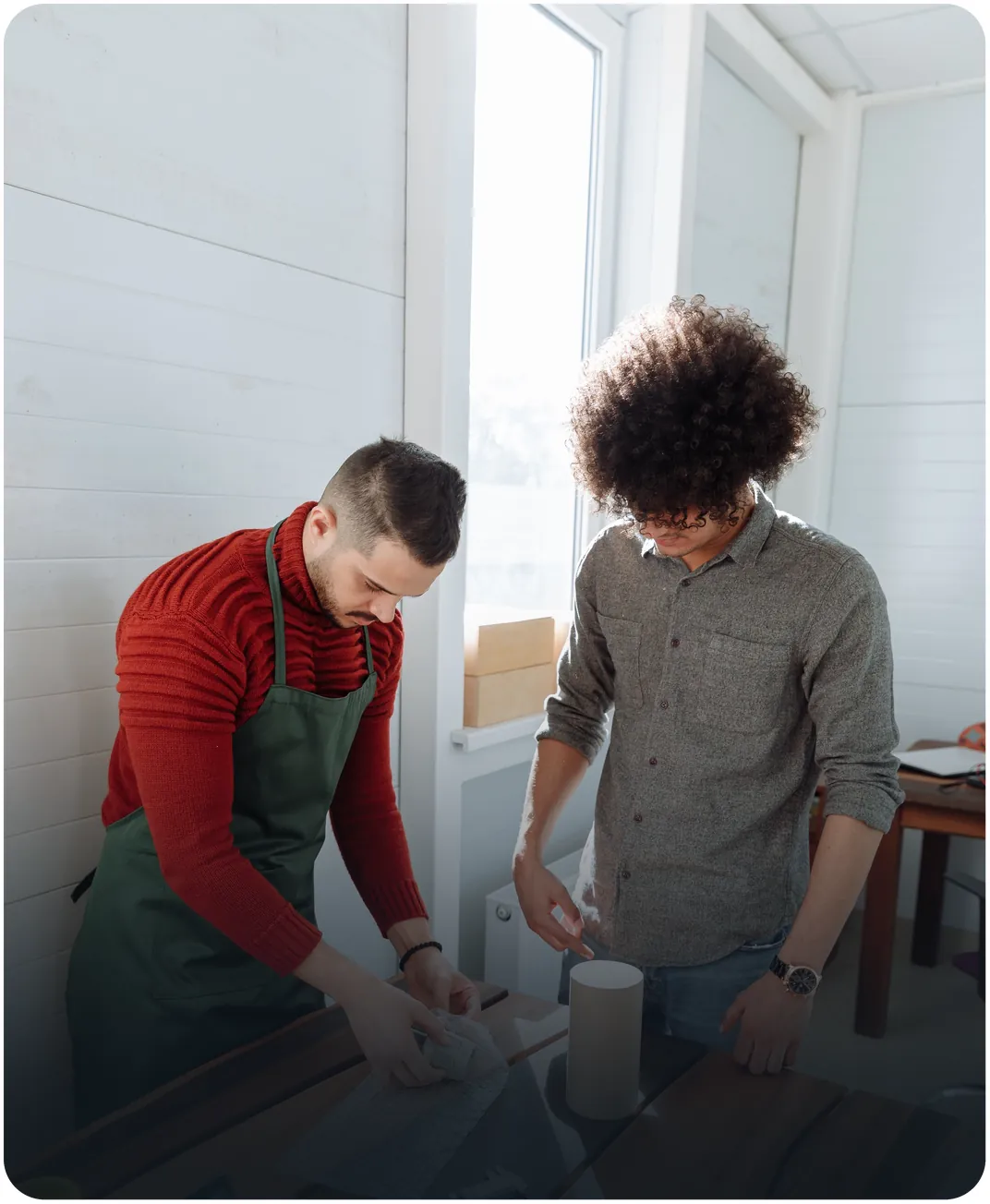 Two men wrapping products at a wooden worktable with cardboard boxes stacked on the windowsill