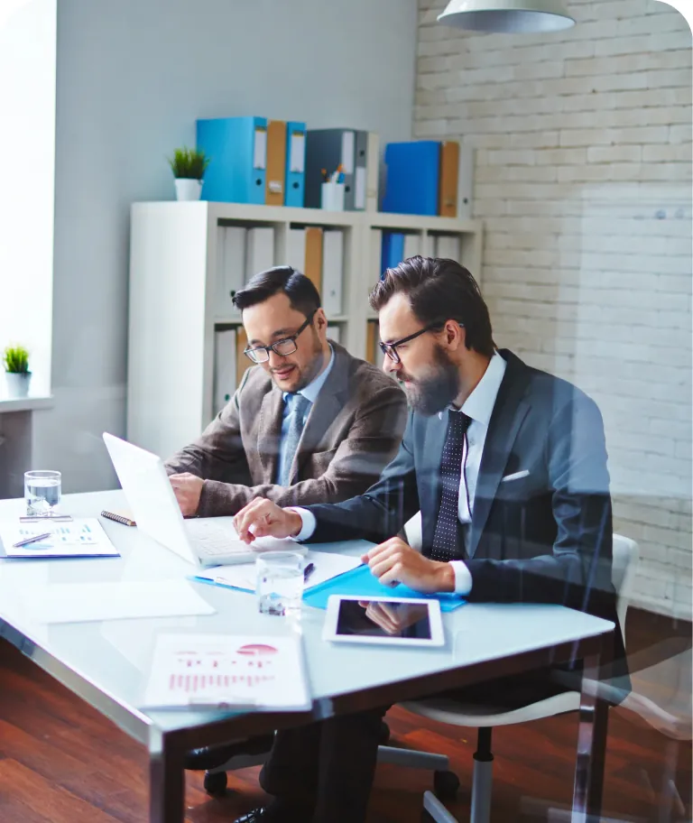 Two businessmen in glasses working together on a laptop at a desk with charts, a tablet and binders behind them