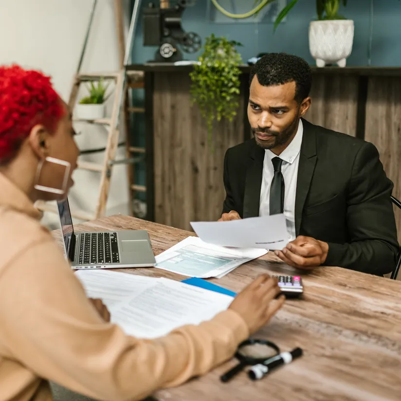 Advisor in a suit reviewing paperwork with a client at a desk with a laptop, calculator and magnifying glass
