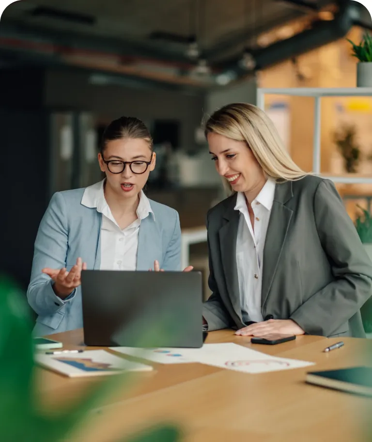 Two businesswomen talking over a laptop at a conference table with printed charts and a phone in front of them