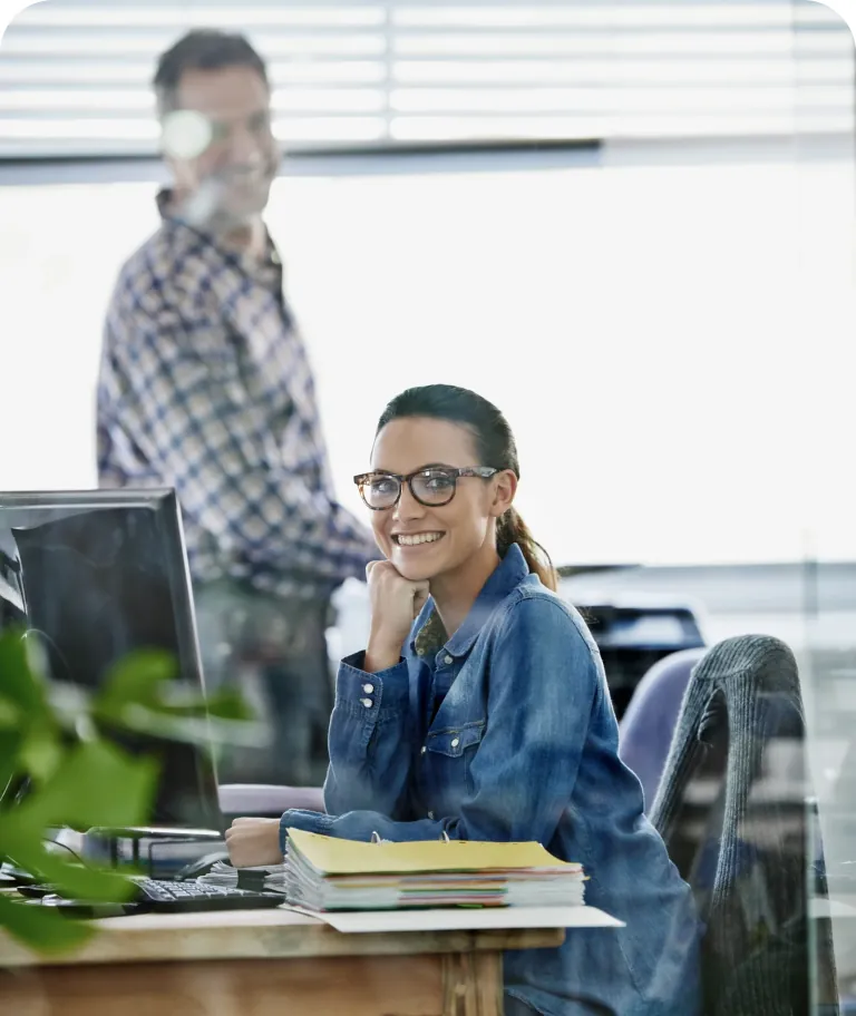 Smiling office worker in glasses at her desk beside a stack of folders, with a colleague standing behind her