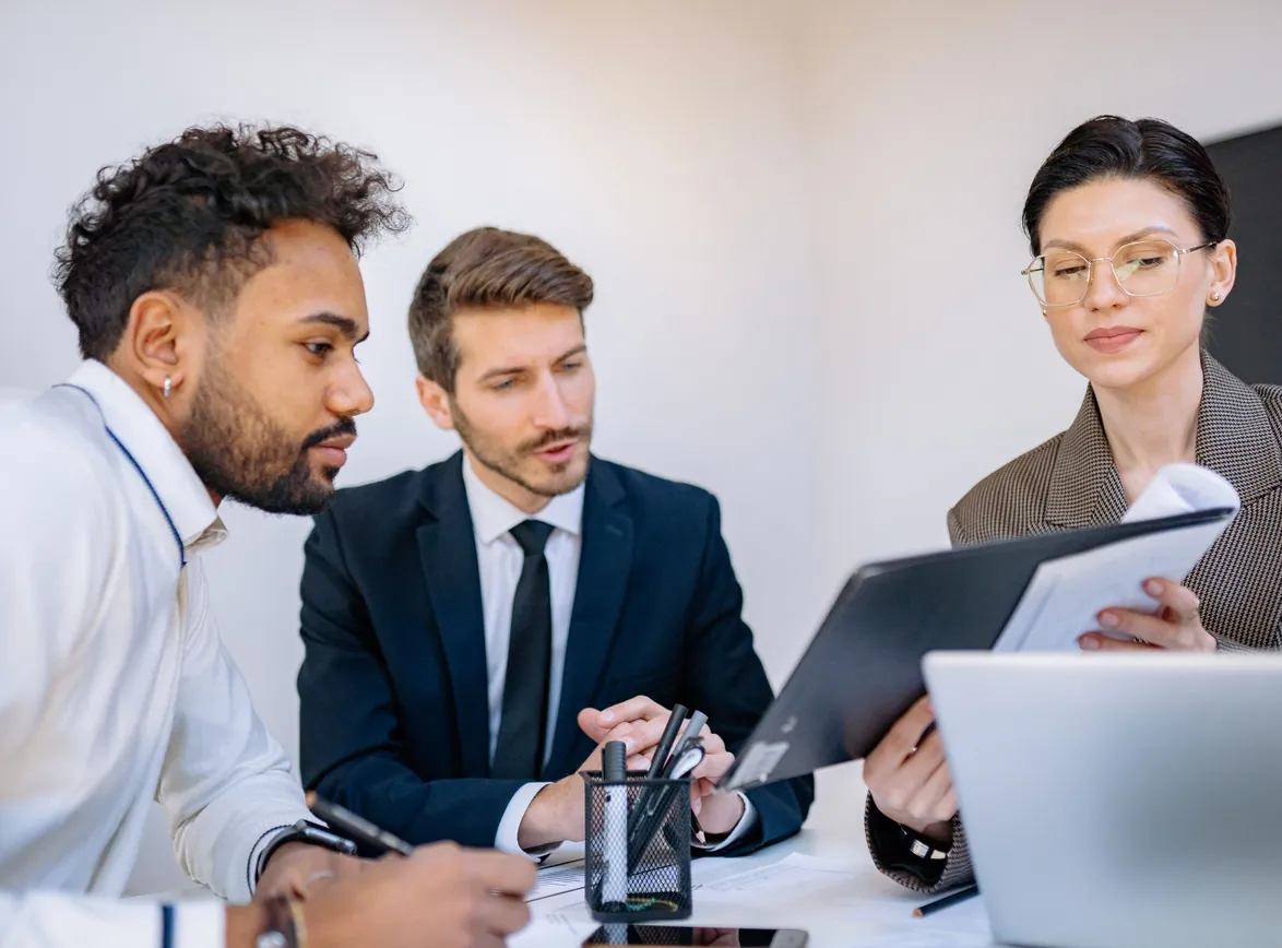 Three professionals look over a clipboard of notes together at an office desk next to a laptop