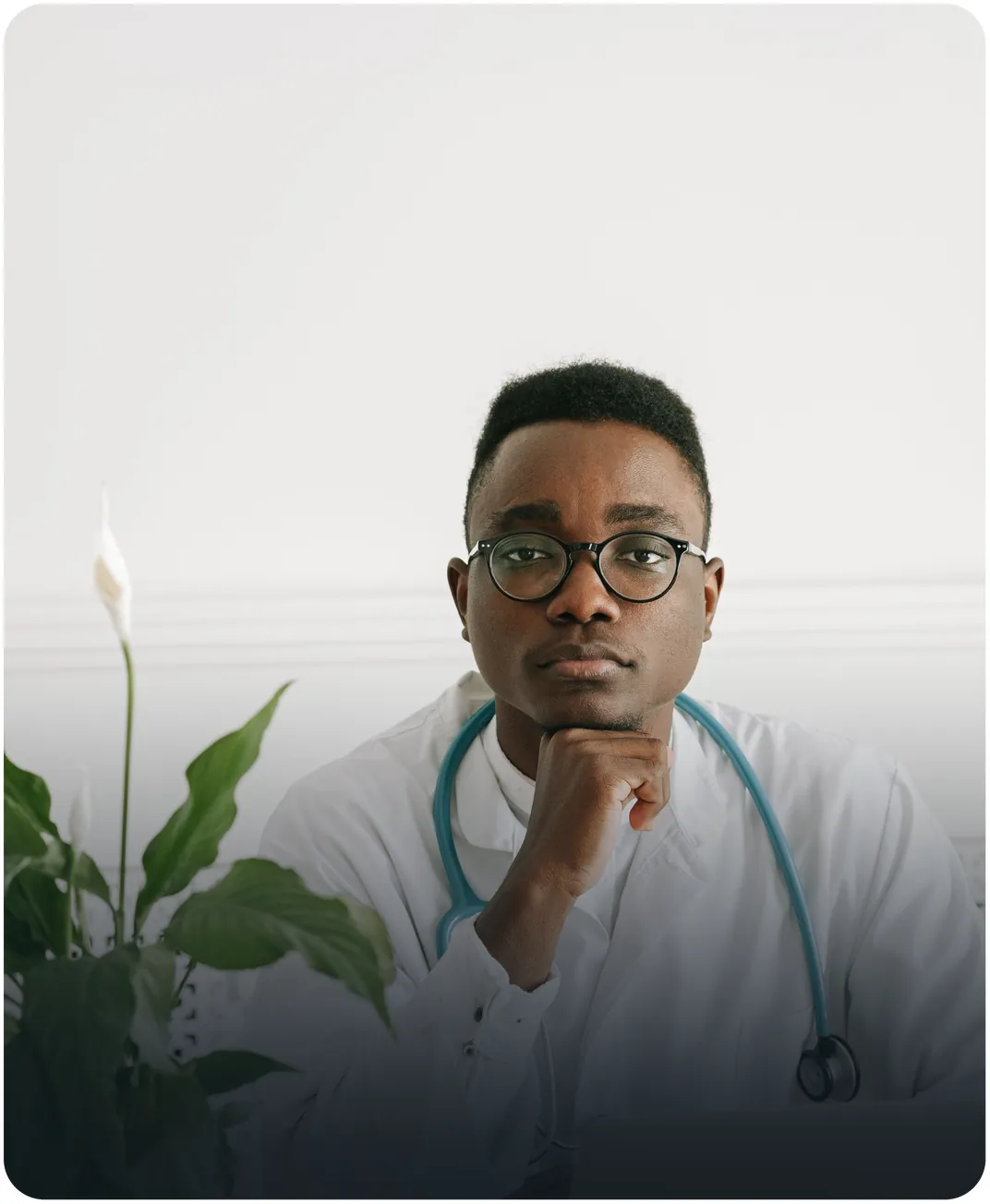Doctor in a white coat with a stethoscope resting his chin on his hand beside a peace lily