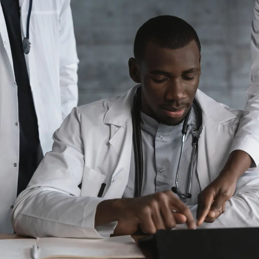 Doctor in a white coat and stethoscope using a tablet at his desk as a colleague points at the screen