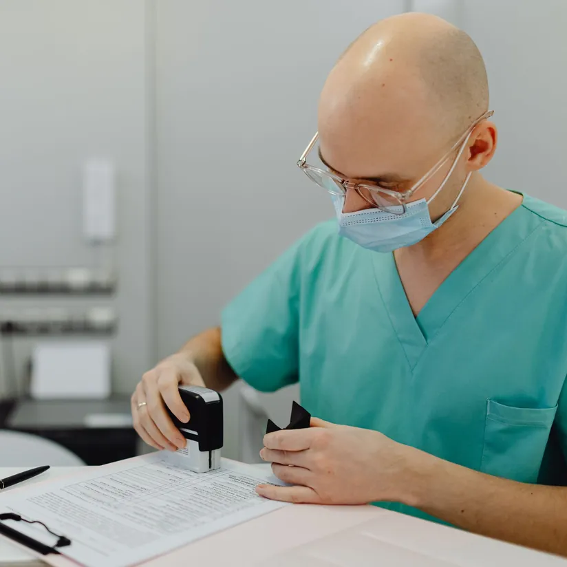 Healthcare worker in teal scrubs and a face mask stamping a printed form at a clinic desk