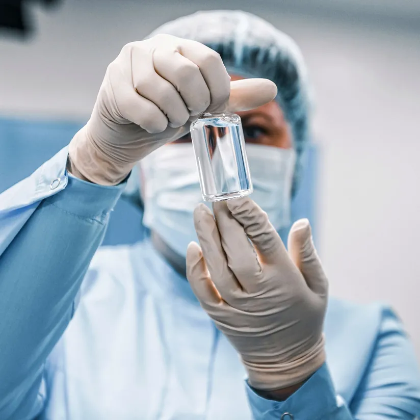 Lab technician in a hairnet, face mask and gloves holding up a glass vial for inspection