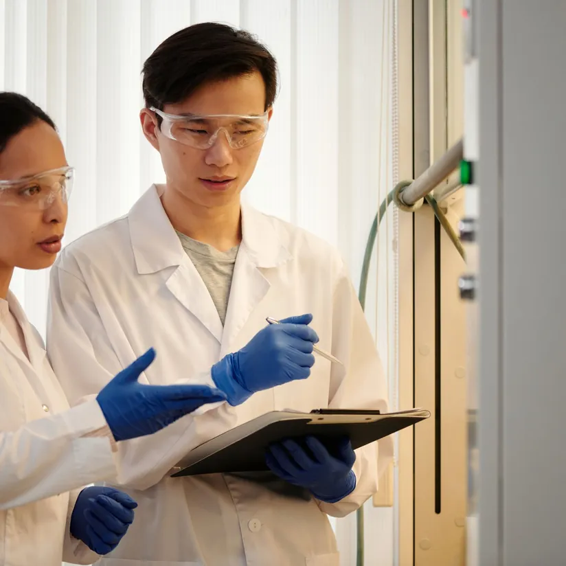 Two scientists in lab coats and safety goggles reviewing notes on a clipboard beside lab equipment