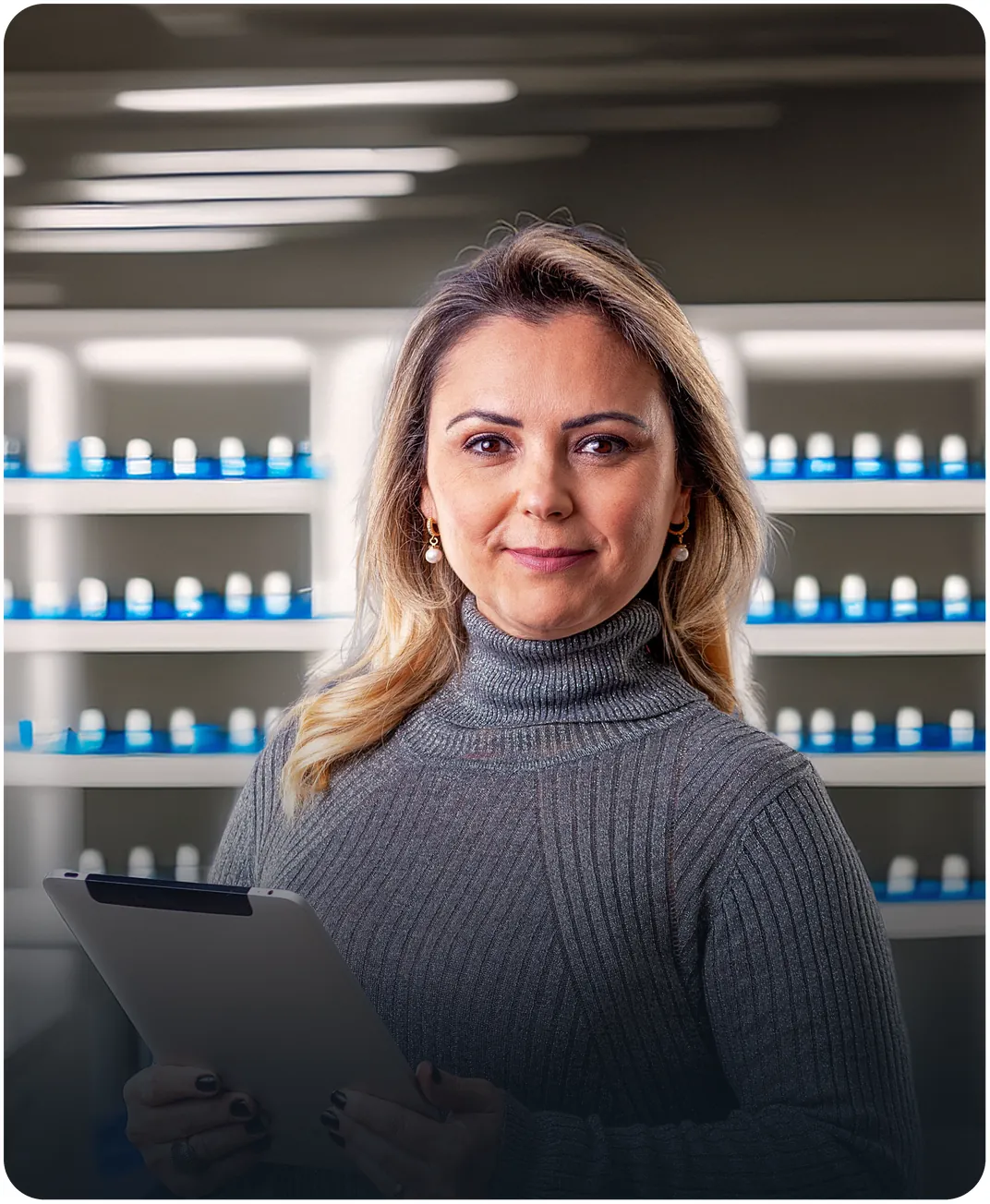 Woman in a gray turtleneck holding a tablet in front of shelves lined with rows of small bottles