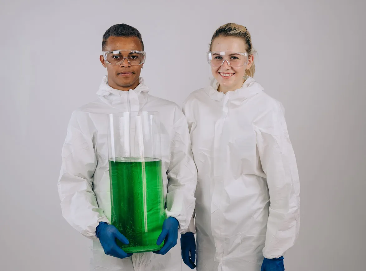 Two scientists in protective gear with a large green chemical vial