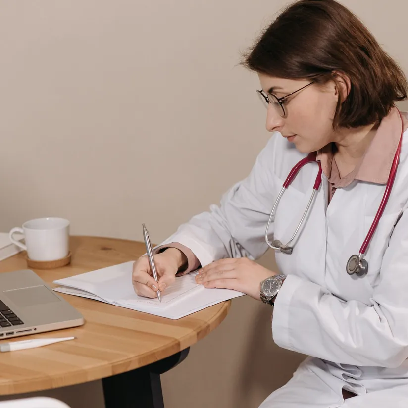 Doctor in a white coat writing notes in a notebook beside a laptop