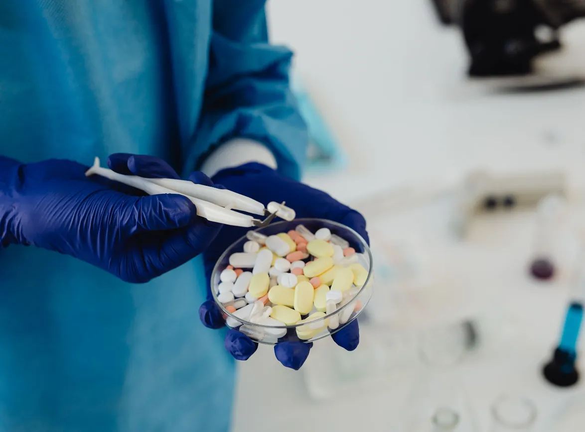 Gloved hands use tweezers to sort tablets and capsules in a petri dish in a lab