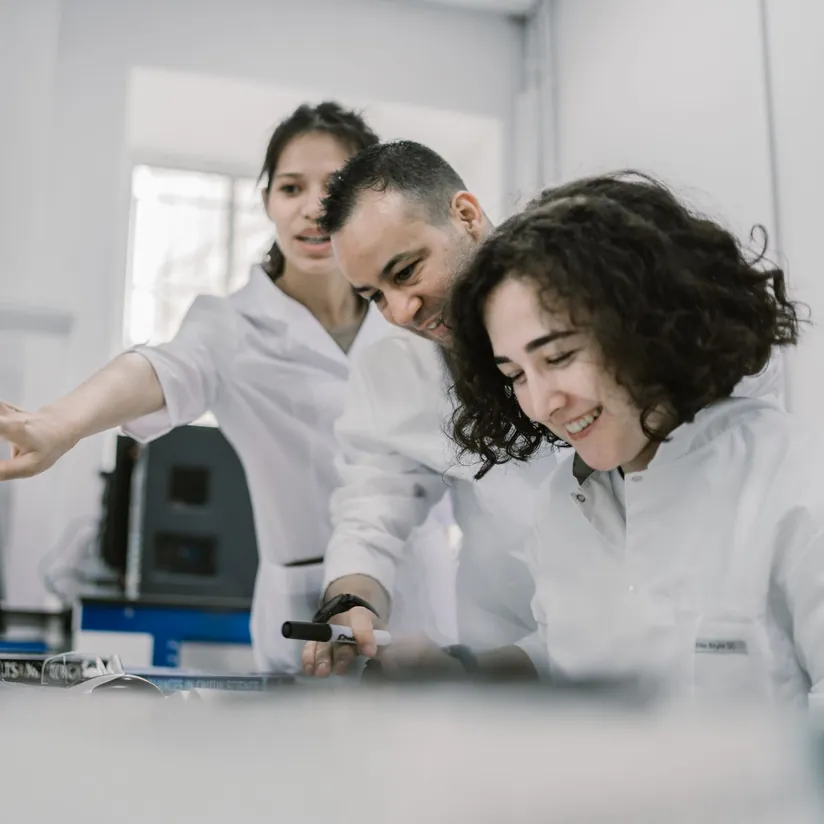 Three scientists in white lab coats leaning over a lab bench, one pointing and one holding a marker