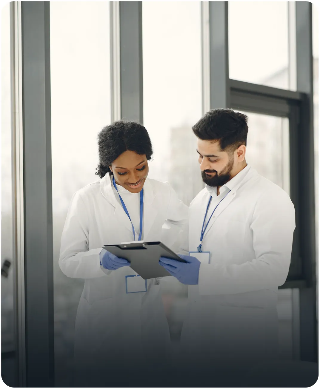 Two scientists in lab coats and blue gloves reviewing notes on a clipboard by a window