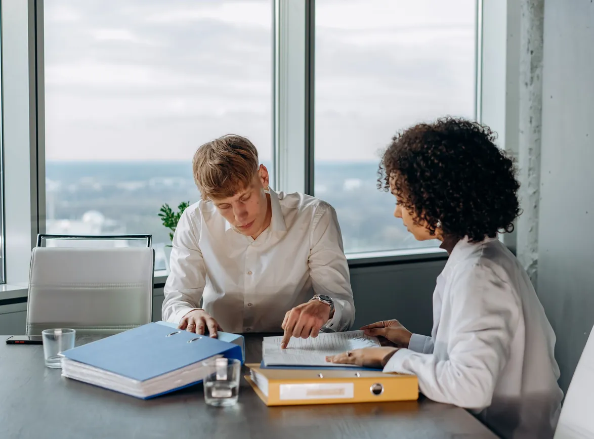 Two colleagues go through a binder of documents together at a table beside a high-rise office window