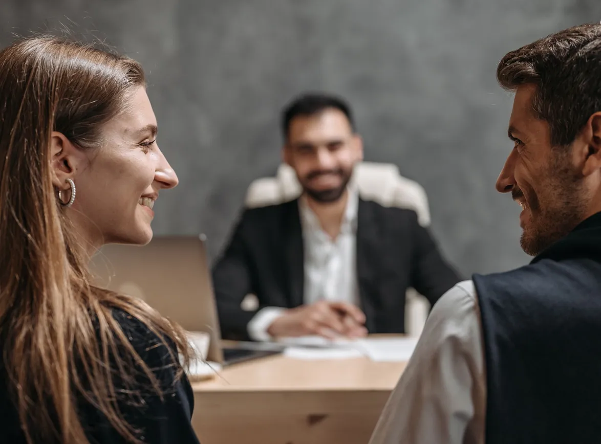 Advisor in a dark suit seated at a desk smiles while talking with two clients sitting across from him