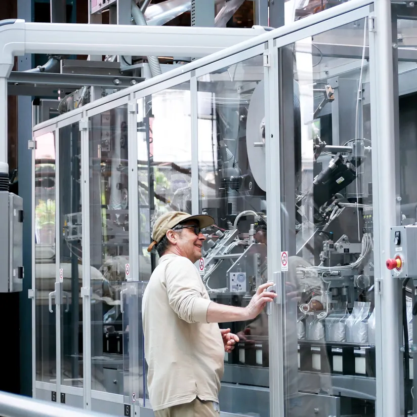 Smiling operator with a hand on the glass enclosure of an automated packaging machine on a factory floor