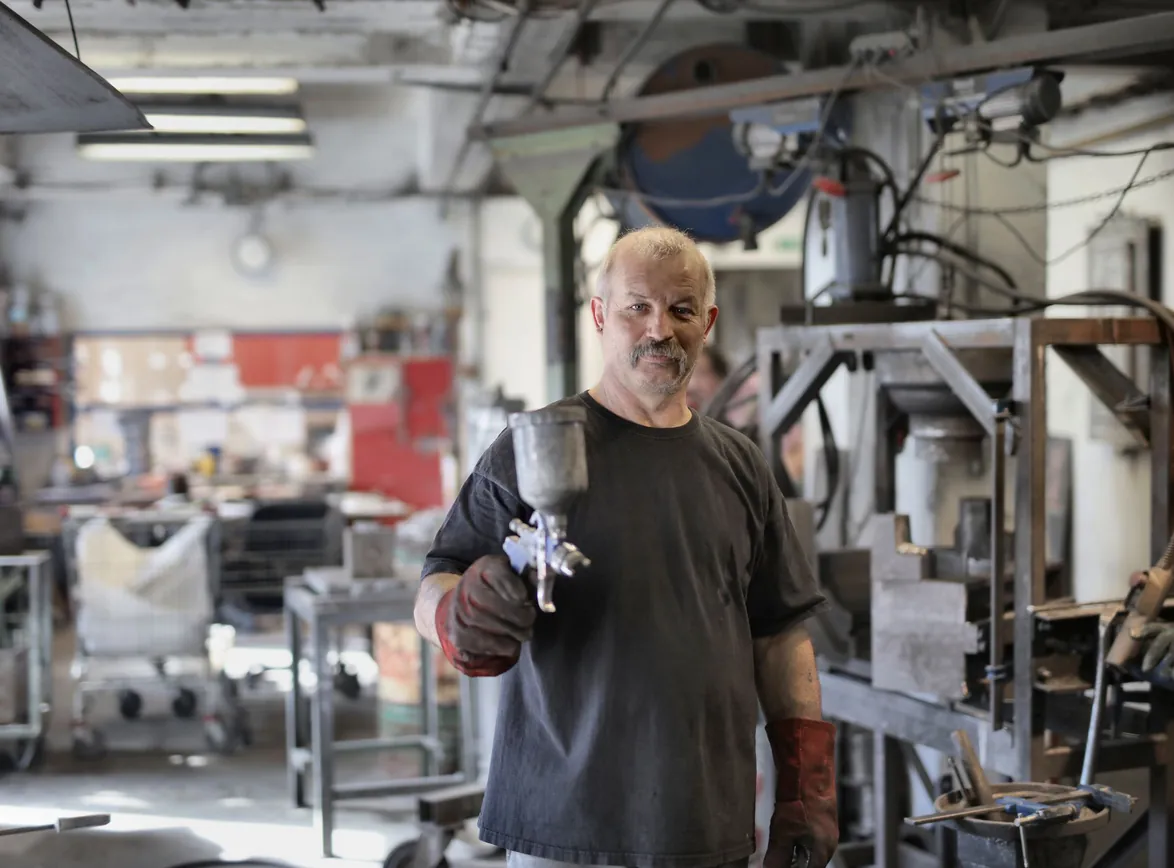 Pleased senior white hair craftsman in heavy duty gloves showing sandblaster and looking at camera