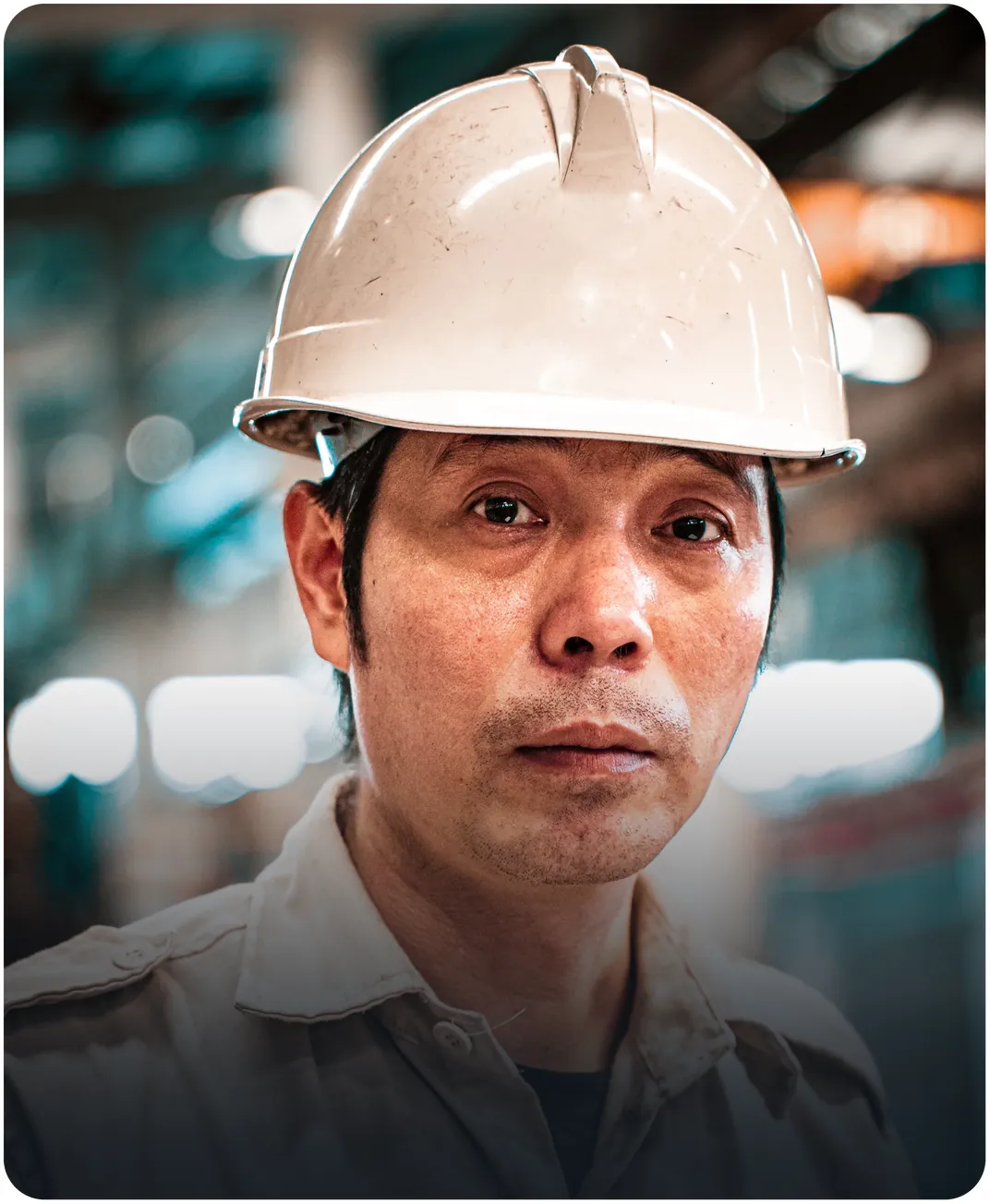 Factory worker in a white hard hat looking straight at the camera on a blurred production floor